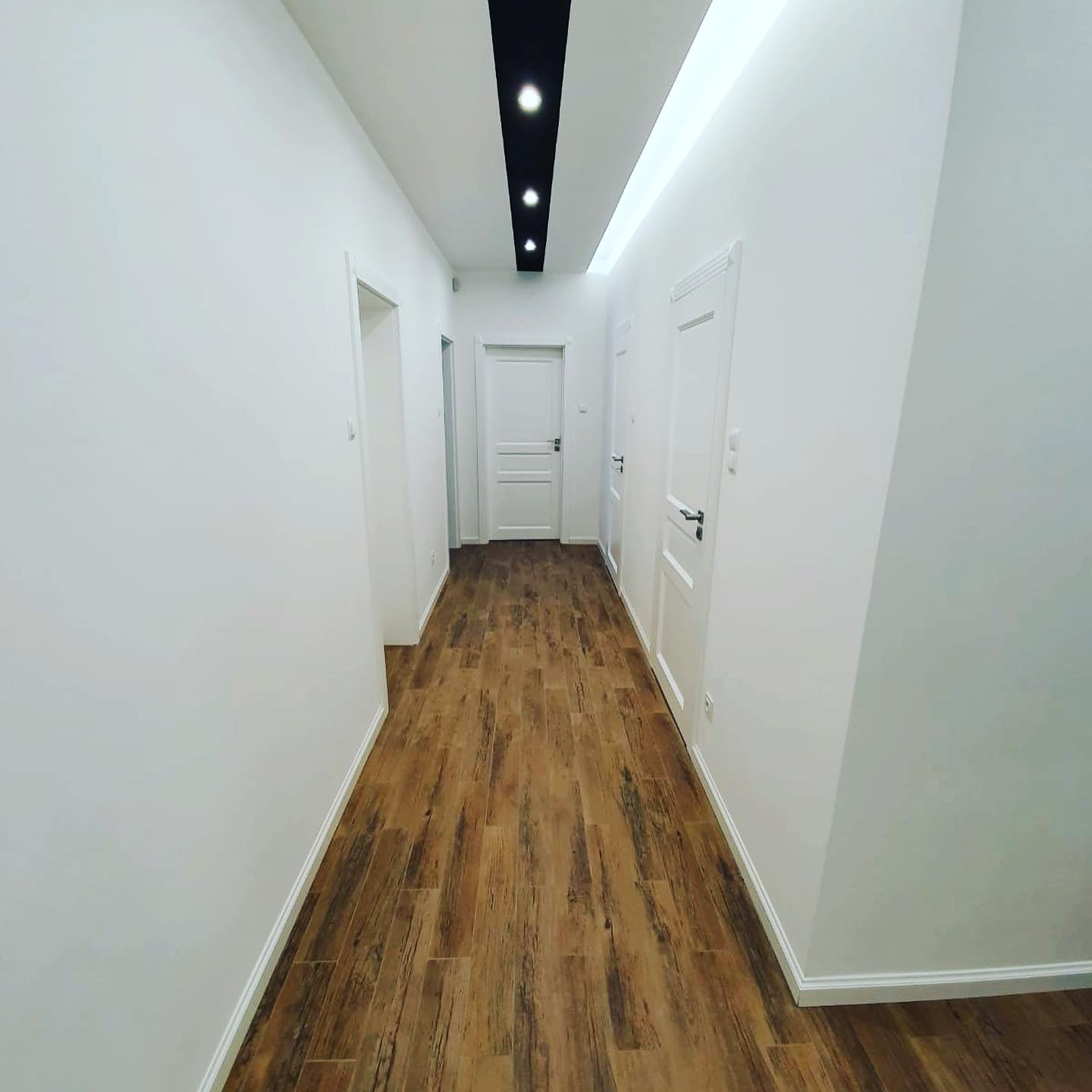 Wypolerowany, jasny korytarz z drewnopodobną podłogą, białymi ścianami i drzwiami, oświetlony listwą LED i czarnym pasem z punktowymi lampami w suficie.