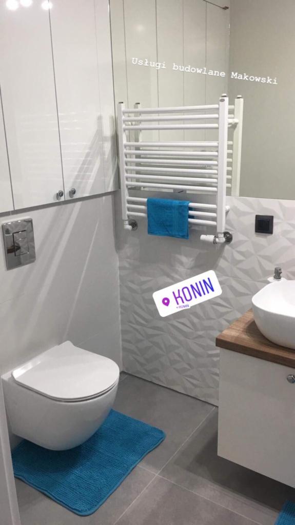 Nowoczesna łazienka po remoncie: podwieszana toaleta, grzejnik drabinkowy z turkusowym ręcznikiem, umywalka na drewnianym blacie, szare płytki podłogowe i dekoracyjne białe płytki ścienne...