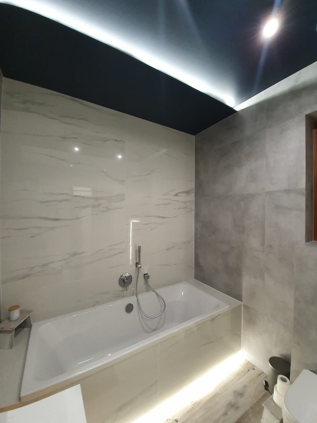 Nowoczesna łazienka z białą wanną, marmurowymi płytkami na ścianach, ciemnym sufitem z oświetleniem LED i subtelnym podświetleniem wanny.