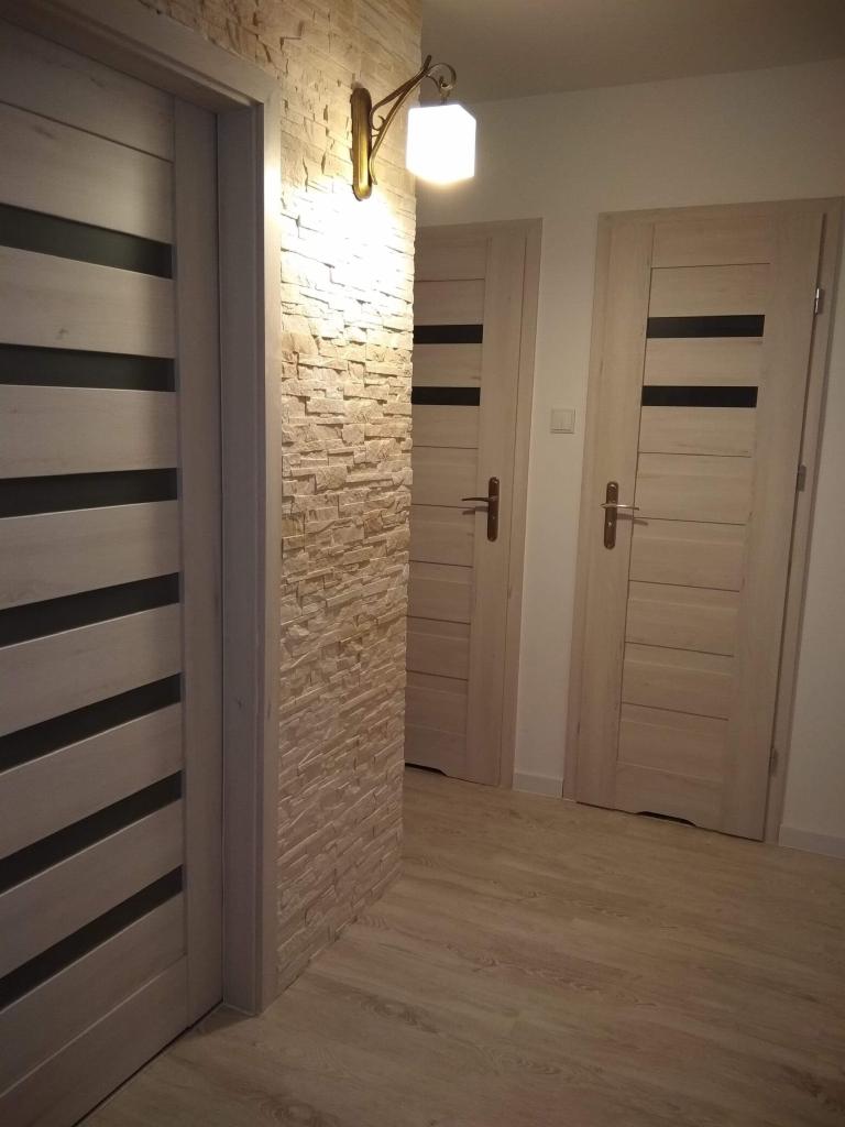 Wnętrze korytarza z trzema drewnianymi drzwiami z czarnymi paskami, ściana wyłożona dekoracyjnym kamieniem i oświetlona kinkietem.