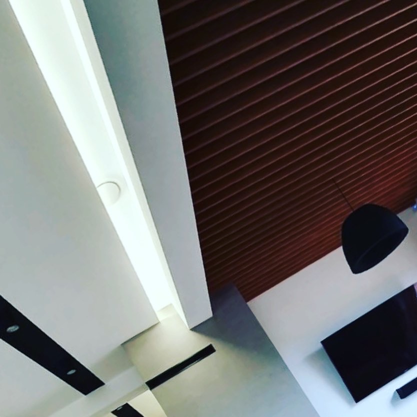 Nowoczesne wnętrze z brązowym, lamelowym sufitem, oświetleniem LED i minimalistyczną lampą wiszącą, widziane z perspektywy ukośnej.