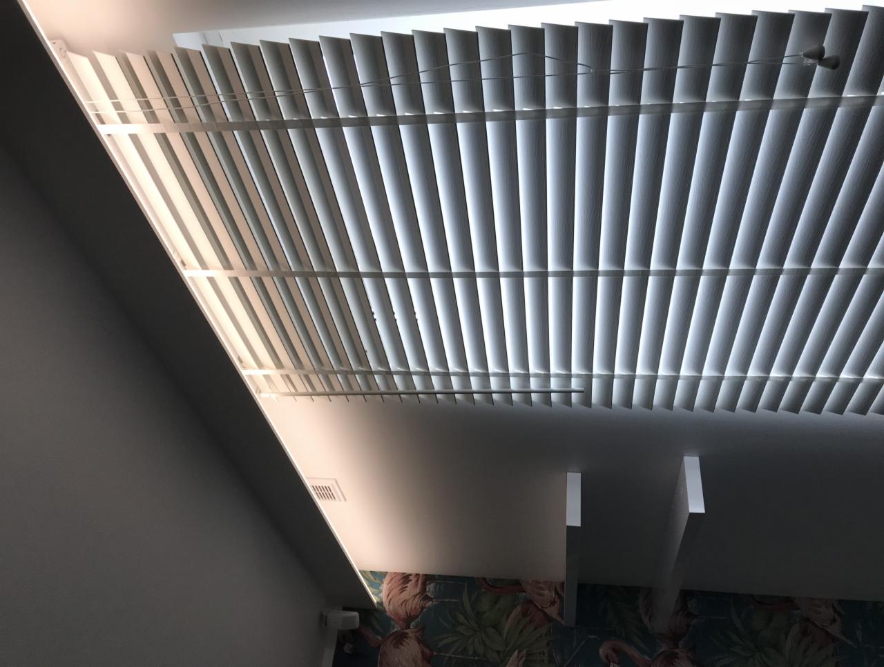 Białe żaluzje pionowe zamontowane pod sufitem, widoczne z dołu, z fragmentem ściany z wzorem w tle.