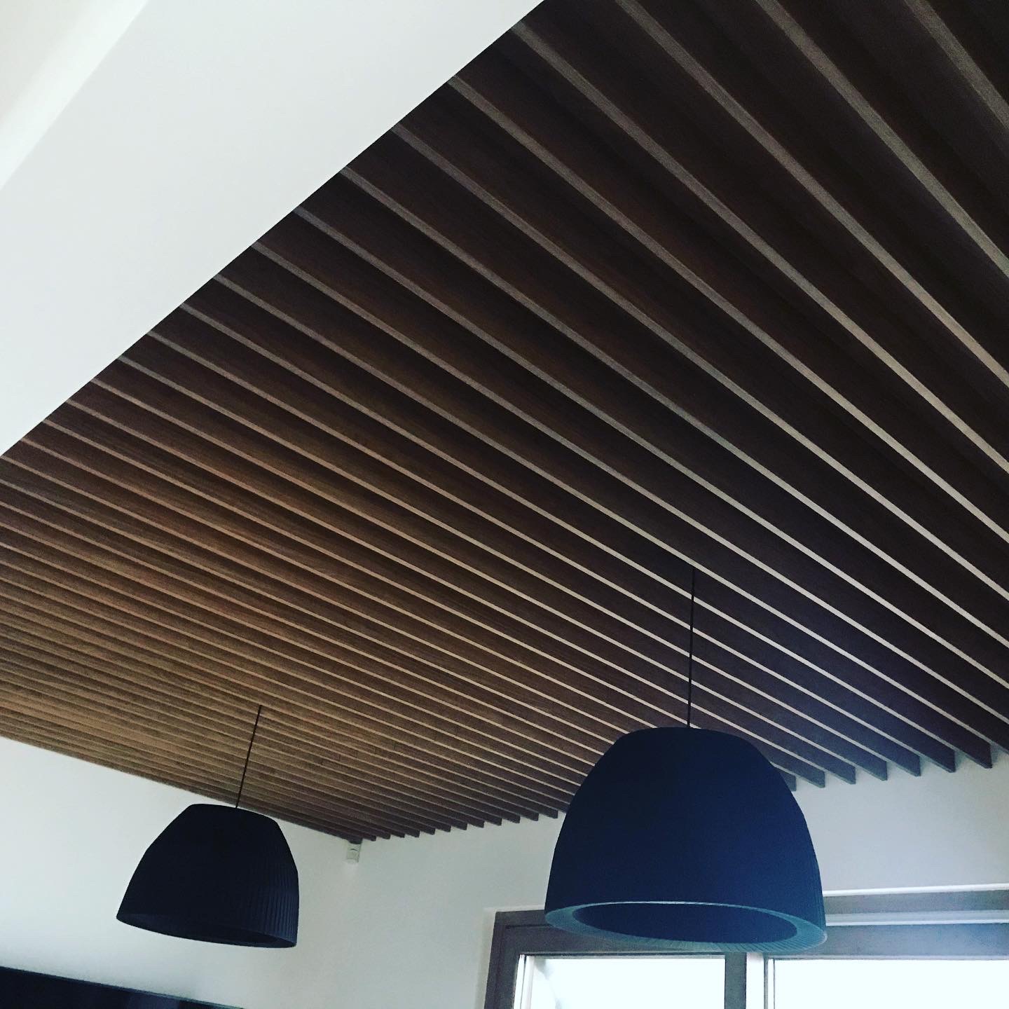 Drewniany sufit lamelowy z równoległymi listwami, oświetlony dwoma czarnymi lampami wiszącymi w kształcie kopuł, widok z dołu.