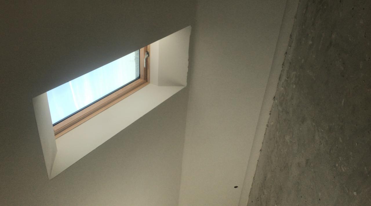 Wnętrze poddasza ze świeżo pomalowanym oknem dachowym, widoczne surowe wykończenie ściany po prawej stronie.