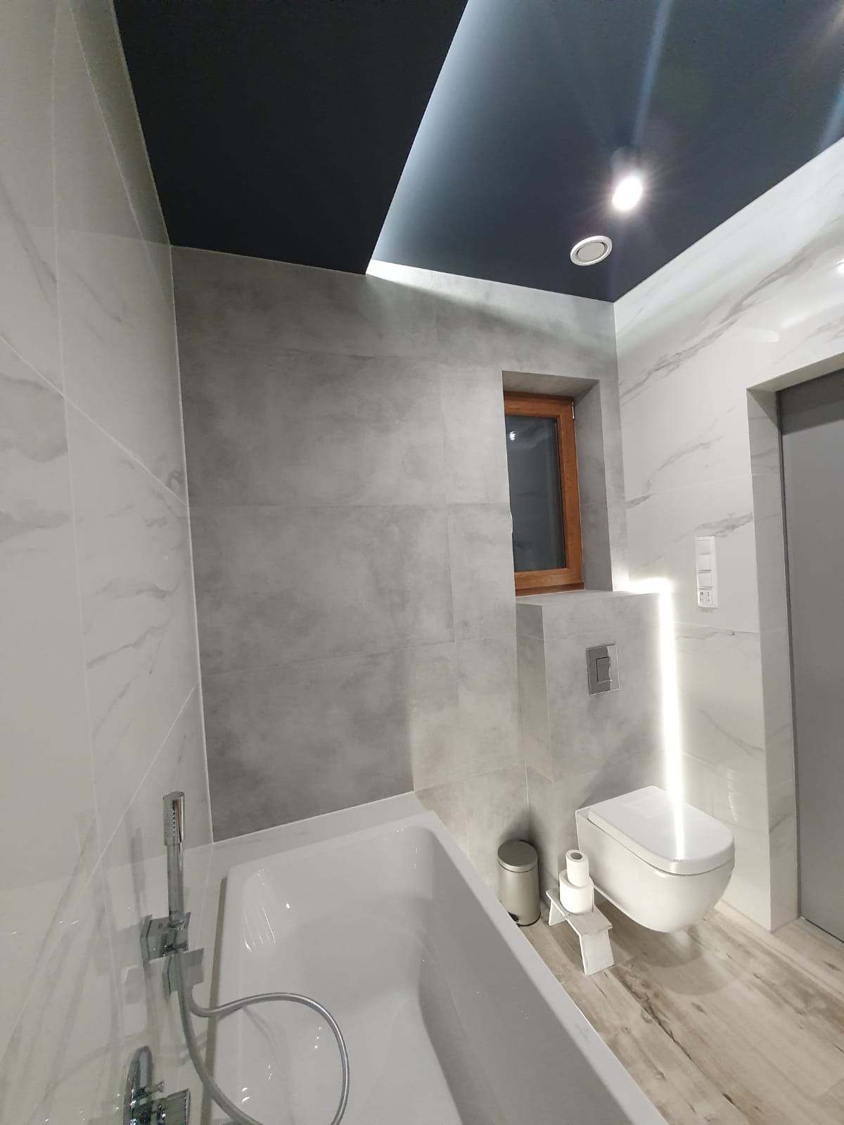Nowoczesna łazienka z wanną, toaletą podwieszaną z podświetleniem LED, szarą mozaiką na ścianach oraz czarnym sufitem podwieszanym z oświetleniem punktowym.