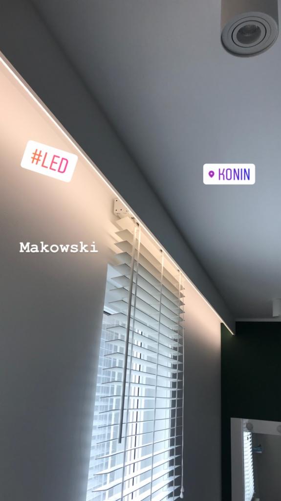 Białe żaluzje w oknie z oświetleniem LED zamontowanym w suficie podwieszanym, widoczny fragment lustra i naklejki z napisem #LED oraz Konin