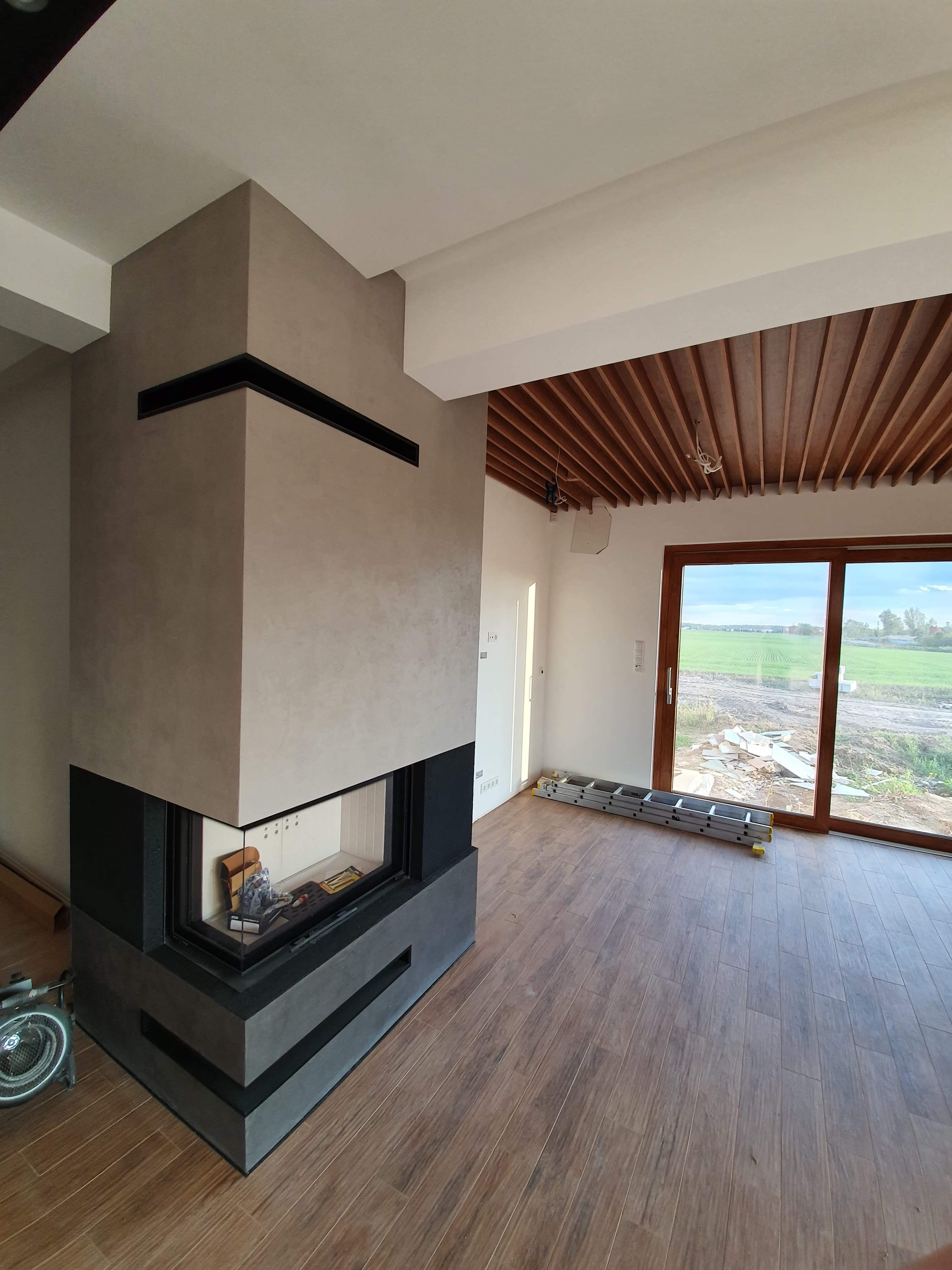 Nowoczesny kominek narożny z szarą obudową i czarnymi elementami, drewniana podłoga, sufit z drewnianymi panelami, widoczne ślady po remoncie i drabina oparta o okno.