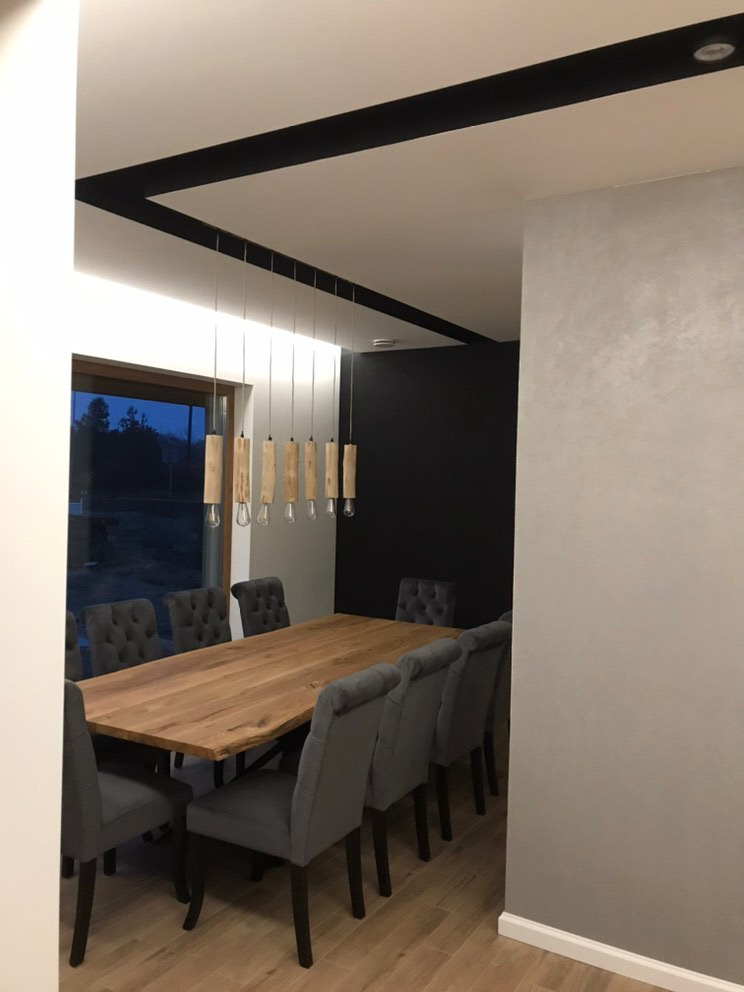 Jadalnia z dużym drewnianym stołem, szarymi krzesłami z pikowaniem i oryginalną lampą z drewnianych elementów, czarna ściana w tle.