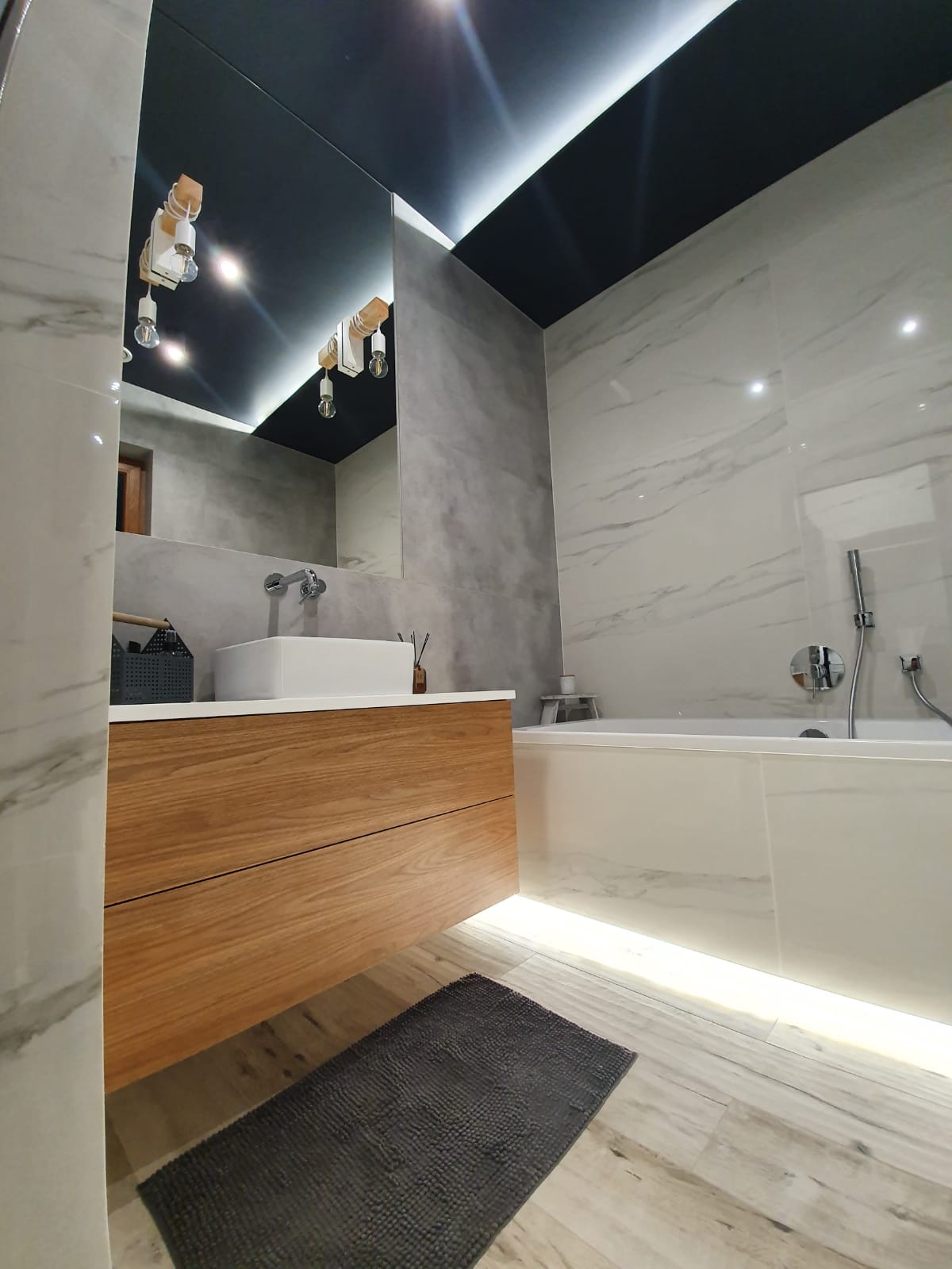 Nowoczesna łazienka z marmurowymi płytkami, drewnianą szafką pod umywalką, czarnym sufitem z oświetleniem LED i szarym dywanikiem podłogowym.
