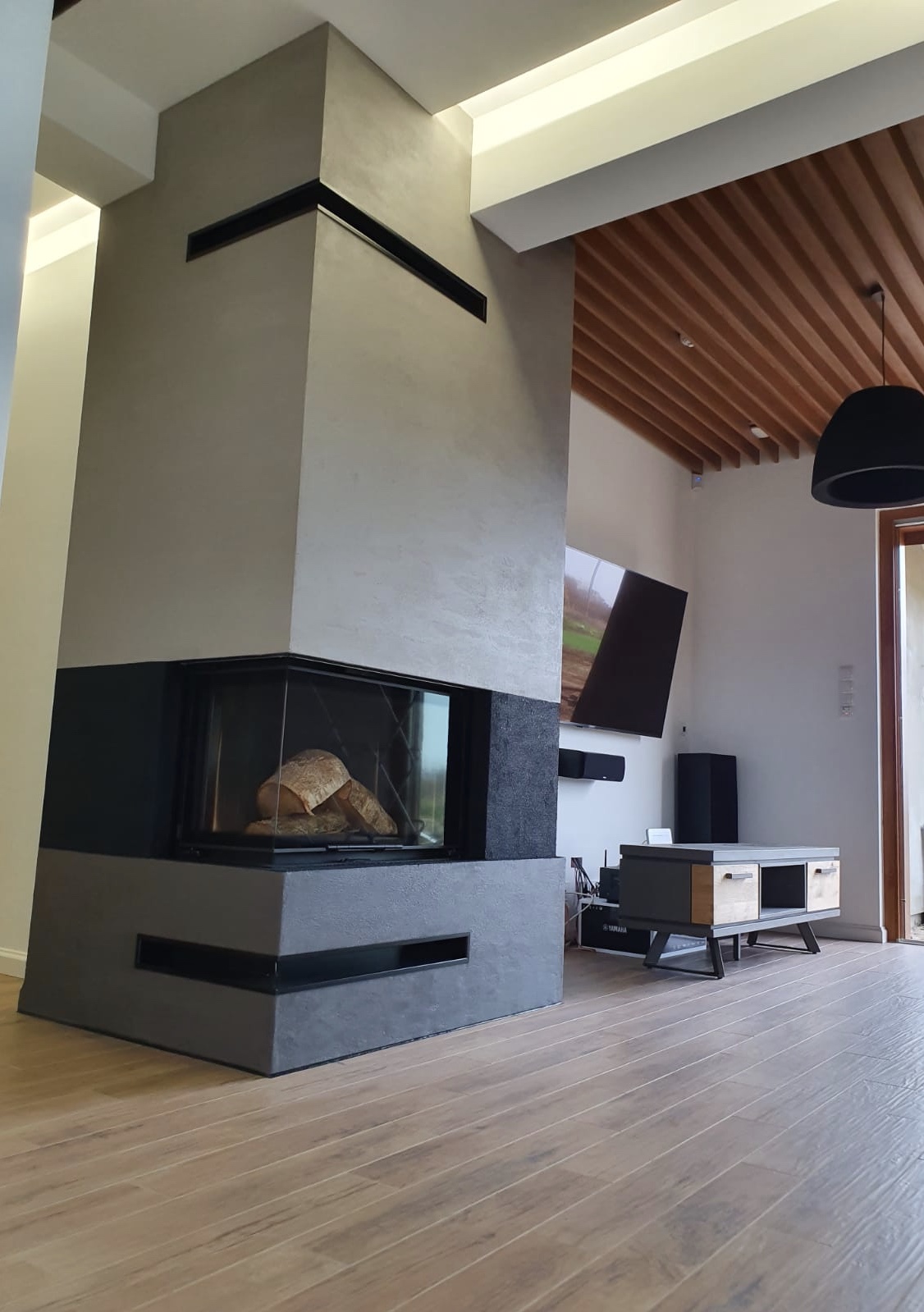 Nowoczesny kominek narożny z obudową w odcieniach szarości i czerni, z drewnem wewnątrz paleniska, na tle jasnego salonu z drewnianym sufitem lamelowym i telewizorem.