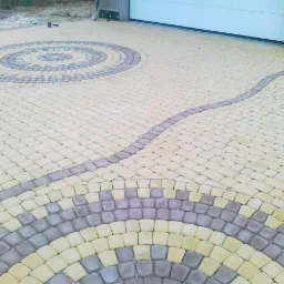 Wzorzysta nawierzchnia z kostki brukowej w odcieniach beżu i szarości, ułożona w okrągłe i łukowe motywy. Widoczny fragment bramy garażowej i narzędzia.