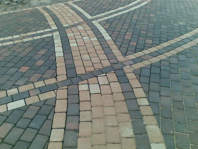 Wzór z kostki brukowej w odcieniach szarości, brązu i beżu, tworzący geometryczny motyw krzyżujących się pasów i łuków.