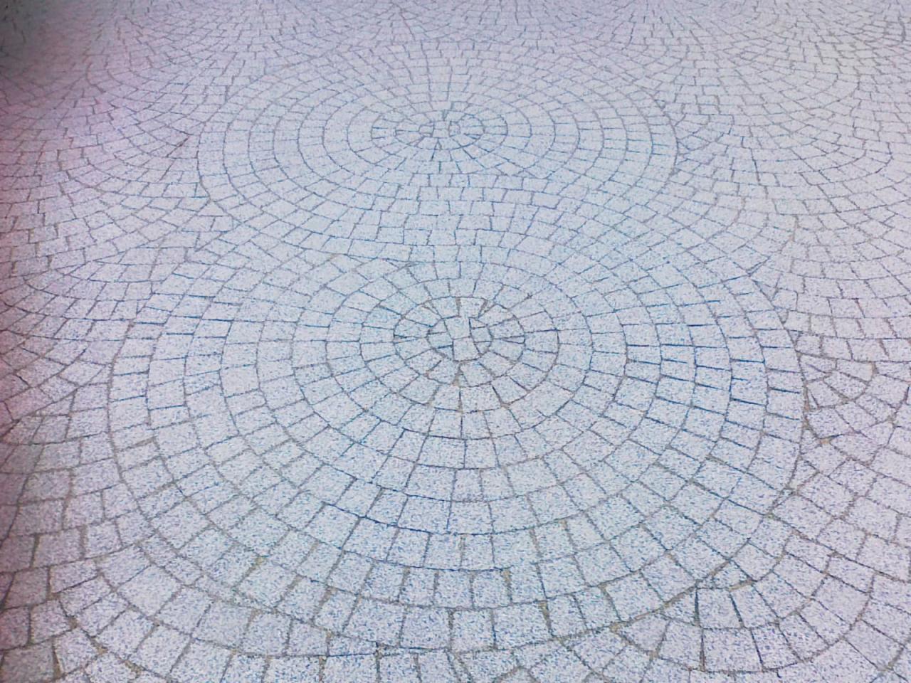 Jasnoszara kostka brukowa ułożona w koncentryczne okręgi, tworząca wzór na placu. Widok z góry, delikatne oświetlenie.