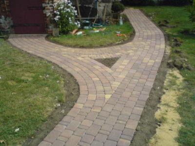 Rozwidlający się chodnik z kostki brukowej, otoczony trawnikiem, z elementami zieleni i kwiatów przy budynku. Widok z perspektywy.