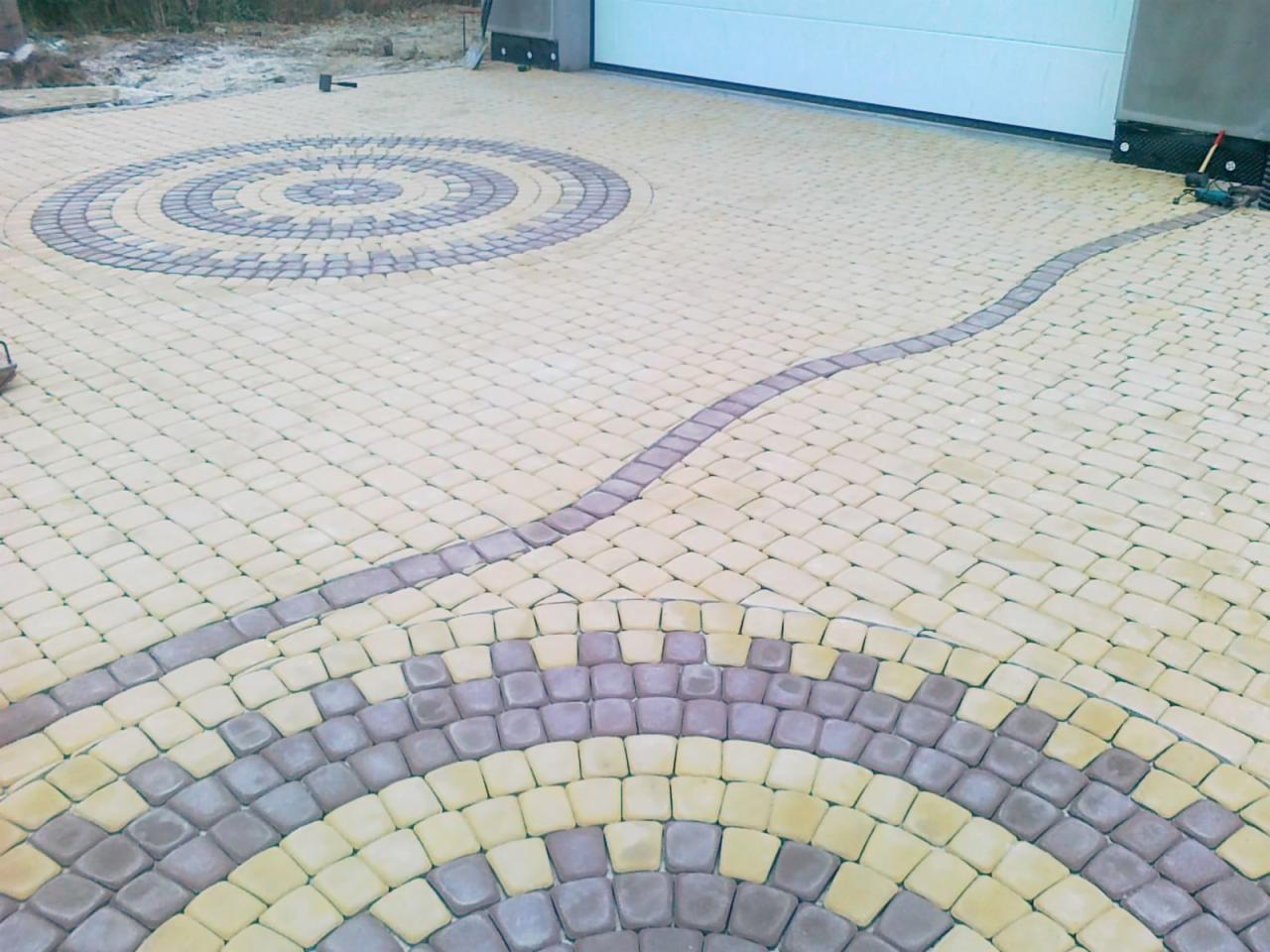 Wzorzysta nawierzchnia z kostki brukowej w odcieniach beżu i szarości, ułożona w okrągłe i łukowe motywy. Widoczny fragment bramy garażowej i narzędzia.