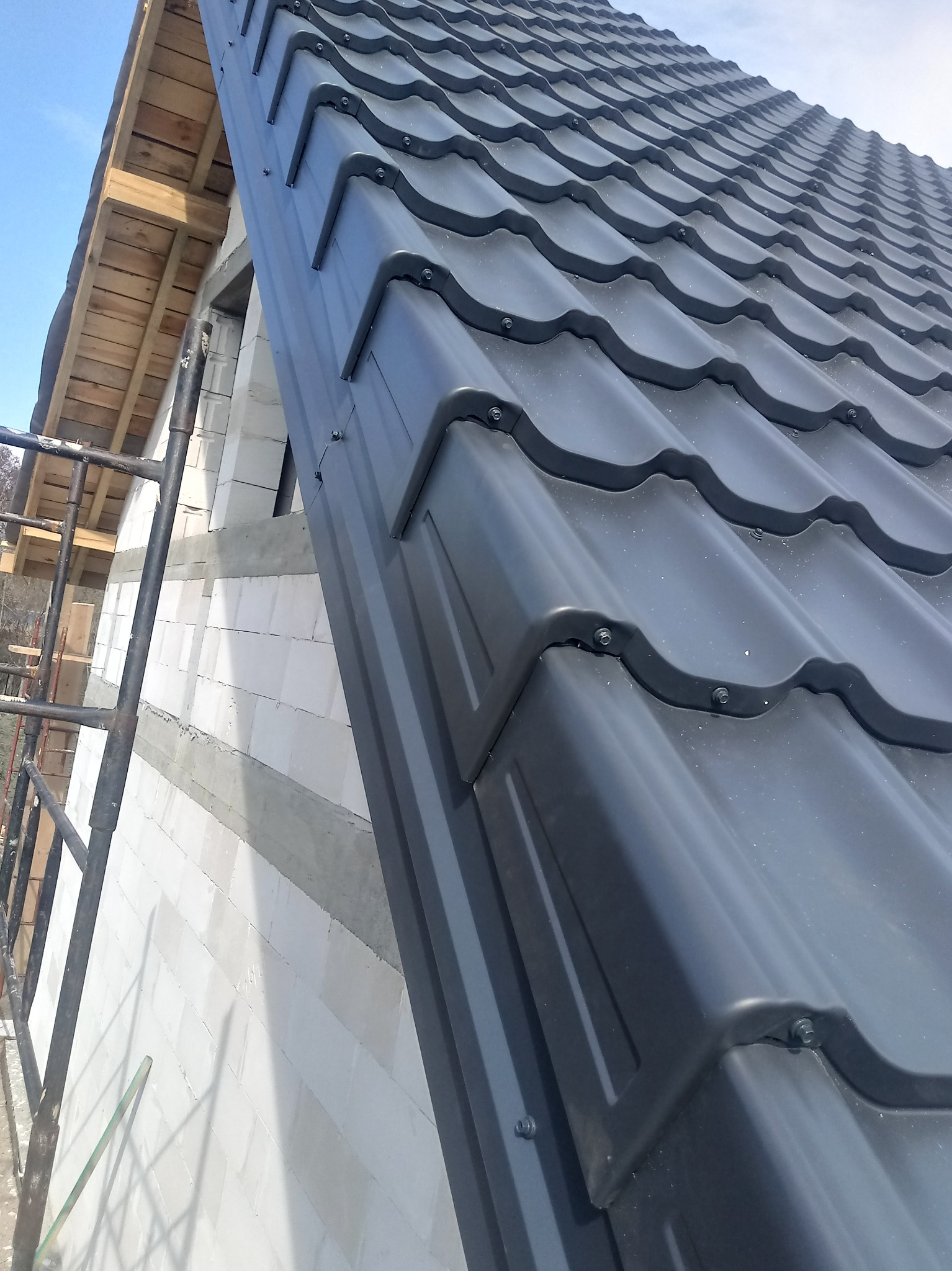 Montaż grafitowej blachodachówki na nowym budynku z białego bloczka, widoczne rusztowanie i drewniana konstrukcja dachu.