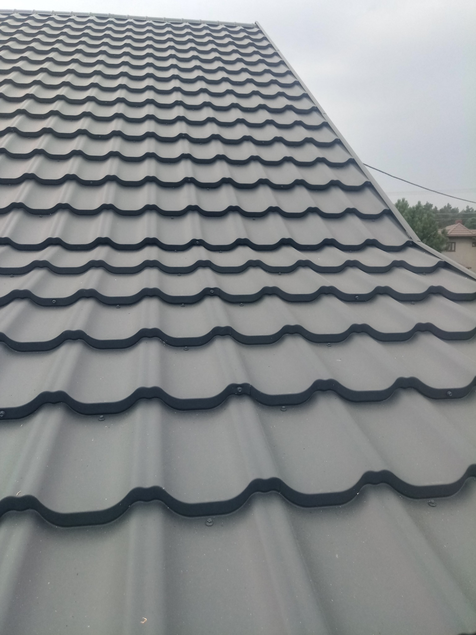 Widok z góry na nowo położone, ciemnoszare blaszane pokrycie dachowe z widocznymi śrubami mocującymi, na tle pochmurnego nieba i fragmentów drzew.