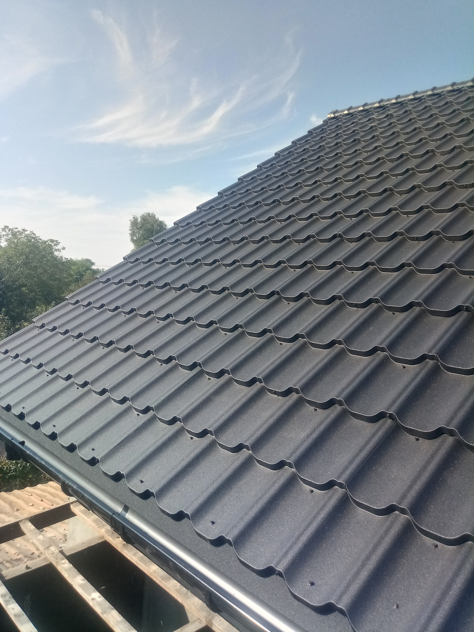 Nowy, ciemnoszary dach z blachodachówki modułowej z zamontowaną rynną, widok z dołu na tle błękitnego nieba z delikatnymi chmurami.