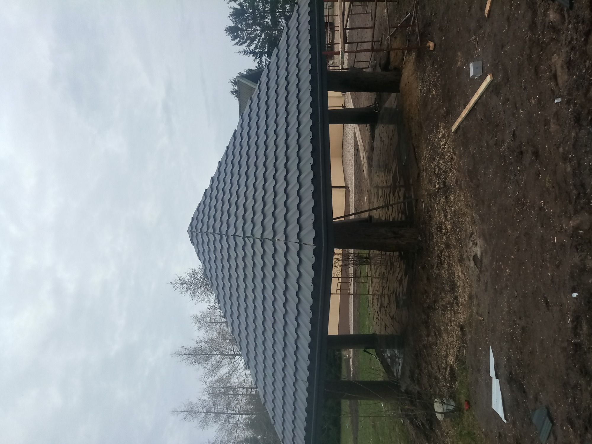 Zadaszona konstrukcja z szarą dachówką karpiówką, widoczna z dołu, na tle pochmurnego nieba i drzew bez liści.