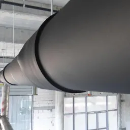 Izolowana rura wentylacyjna, zabezpieczona materiałem armaflex, zawieszona pod betonowym sufitem w surowym wnętrzu budynku w trakcie budowy.