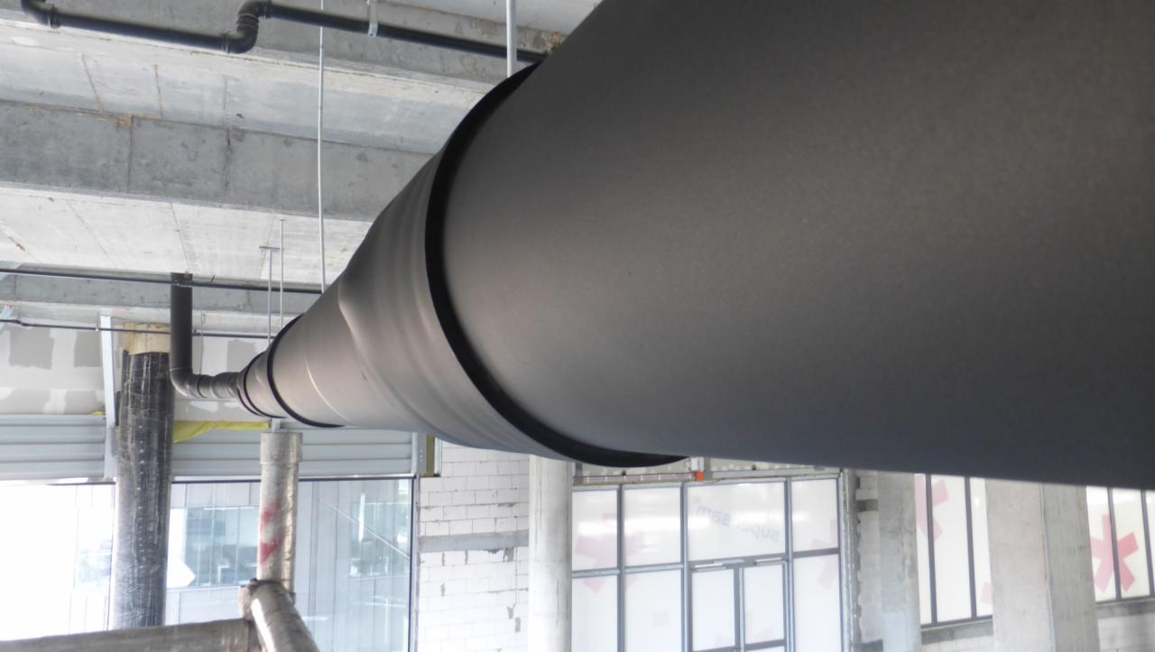 Izolowana rura wentylacyjna, zabezpieczona materiałem armaflex, zawieszona pod betonowym sufitem w surowym wnętrzu budynku w trakcie budowy.