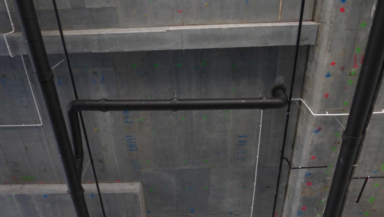 Rurociąg z czarnych rur w surowym, betonowym wnętrzu, z widocznymi kolorowymi oznaczeniami na suficie i ścianach.