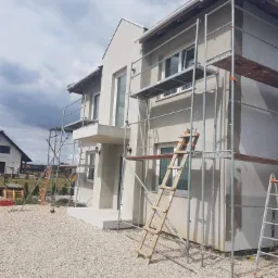 Nowoczesny dom w trakcie tynkowania z użyciem rusztowań, widok z boku na elewację w jasnych barwach, drabiny opierają się o ścianę.