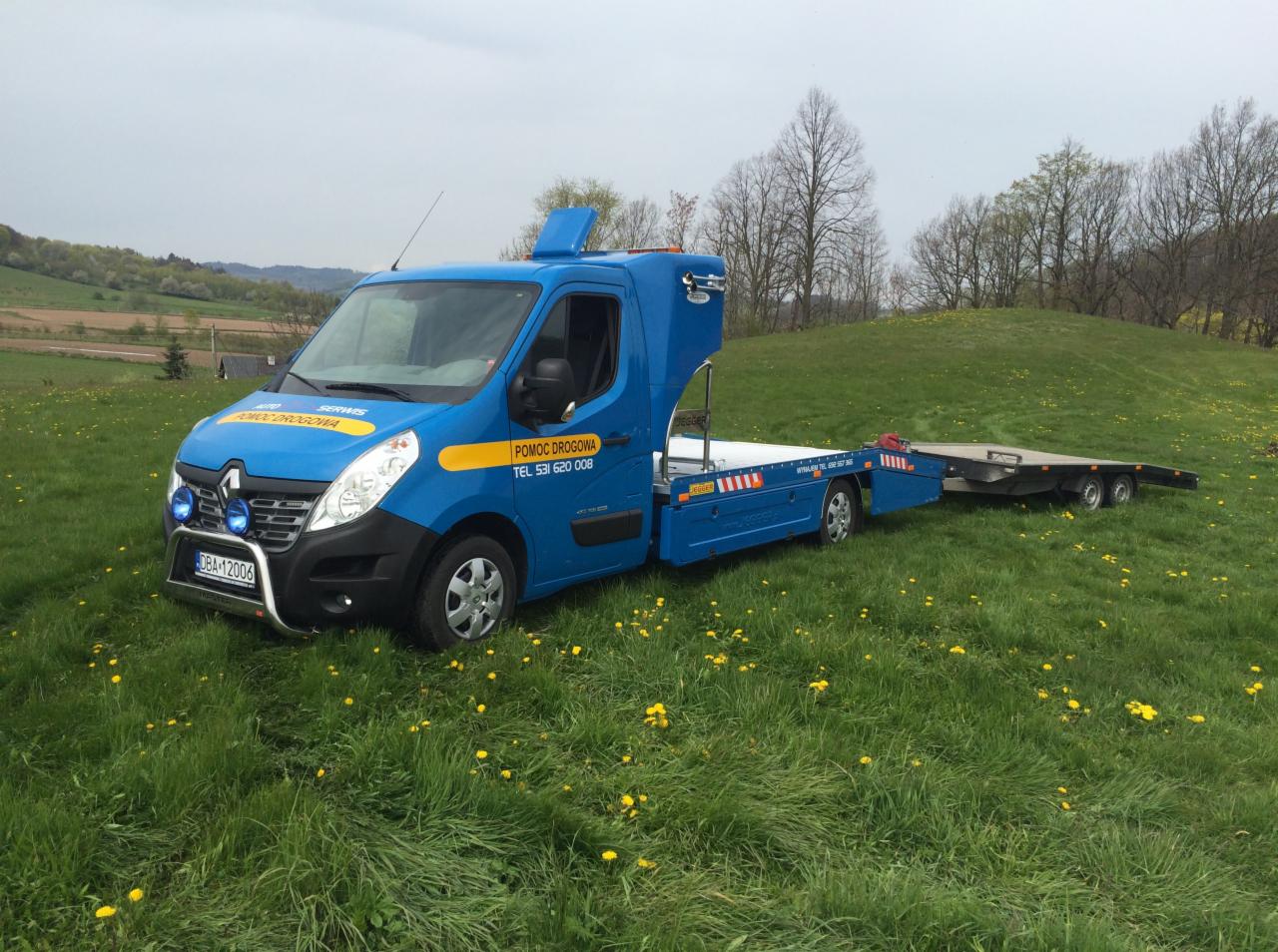 Niebieska laweta z przyczepą stoi na zielonym trawniku z żółtymi kwiatami, w tle pagórkowaty krajobraz.