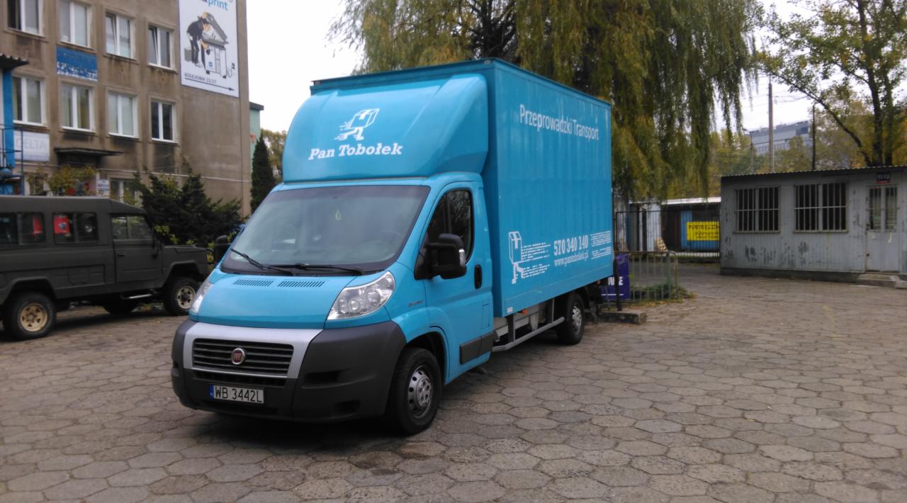 Niebieski bus dostawczy 'Pan Tobołek' z napisem 'Przeprowadzki Transport' na tle budynku i kostki brukowej. Widoczny numer telefonu i adres strony internetowej.
