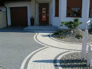 Elegancki podjazd z kostki brukowej w odcieniach szarości i beżu, z dekoracyjnymi obrzeżami i roślinami, prowadzący do domu z garażem i ciemnymi drzwiami.