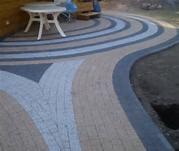 Falista ścieżka z kostki brukowej w odcieniach beżu, szarości i czerni. Widoczny fragment białego stolika i niezabudowany fragment ziemi. Nietypowy wzór.