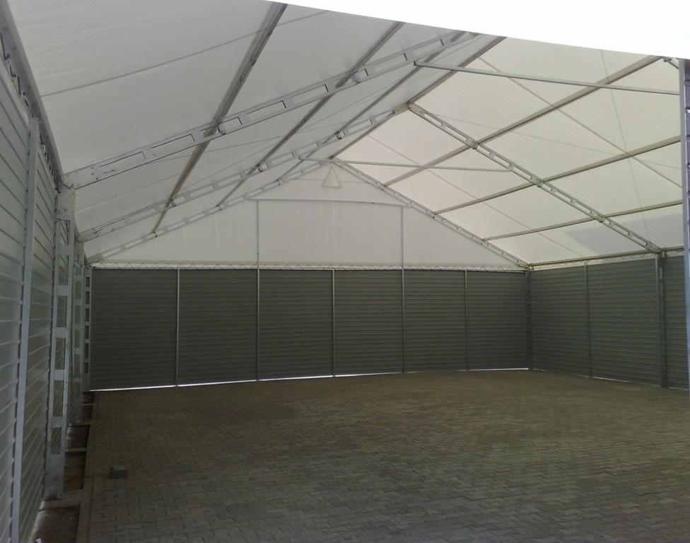 Wnętrze hali z szarą podłogą z kostki brukowej i ścianami z paneli. Widoczna metalowa konstrukcja dachu pokryta białym materiałem. Hala oświetlona naturalnym światłem.