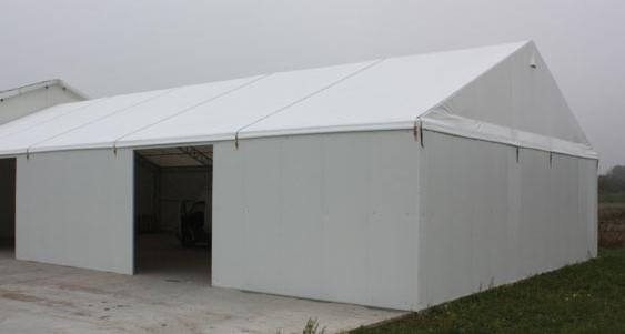 Biała hala namiotowa z płyt warstwowych, widok z zewnątrz, z otwartymi wrotami wjazdowymi. Lekka mgła w tle, betonowa podstawa.