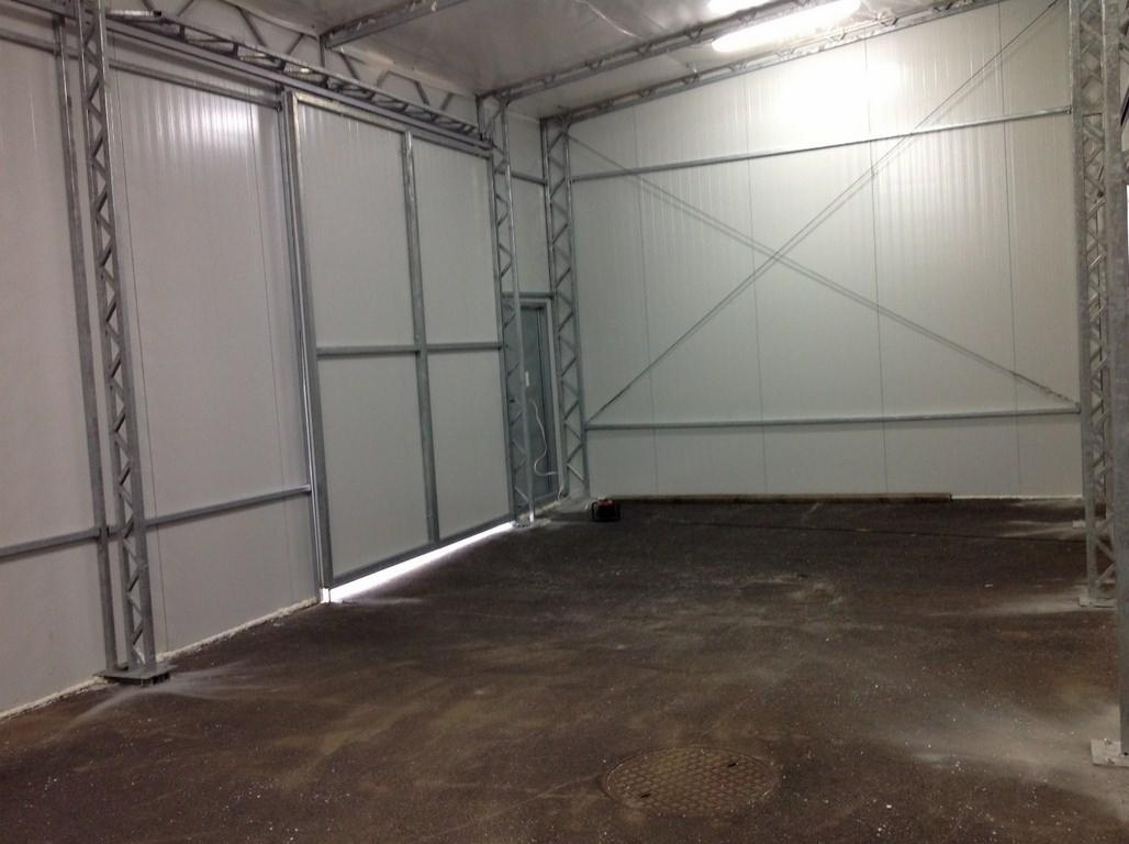 Wnętrze hali z płyt warstwowych, widoczne stalowe konstrukcje nośne i fragment otwartych wrót. Podłoga betonowa z widoczną studzienką kanalizacyjną.