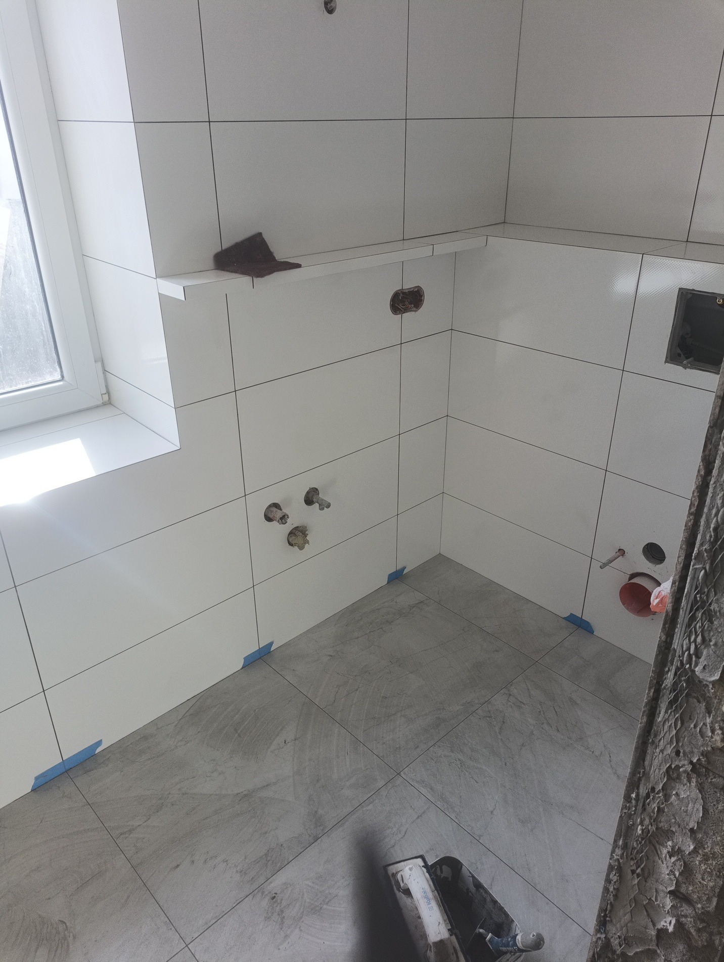 Białe, kwadratowe płytki ścienne i szare podłogowe w trakcie układania. Widoczne niebieskie taśmy maskujące i narzędzia glazurnicze. Półka z płytki pod oknem.