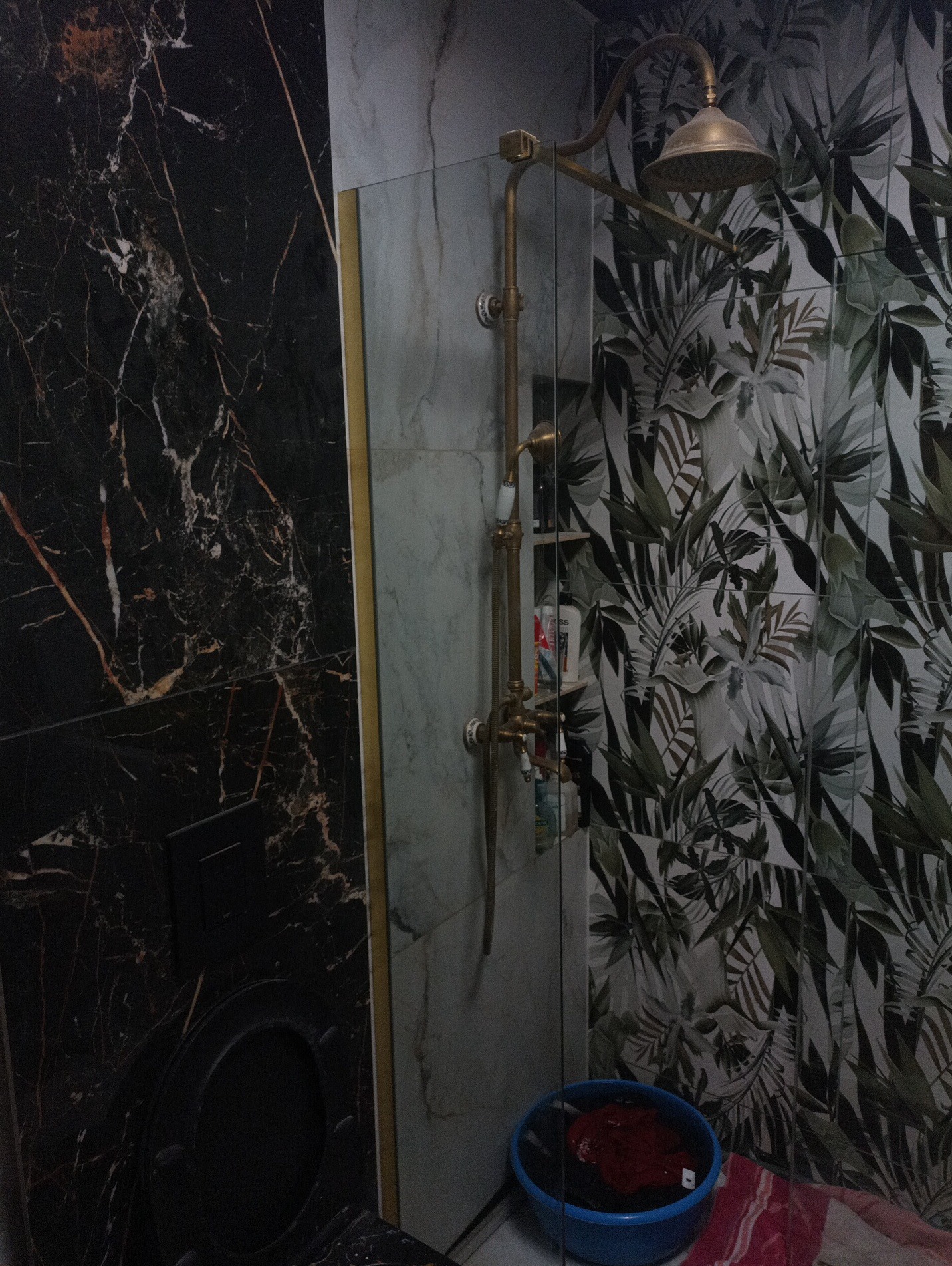 Wykończona łazienka z czarnym marmurem, szklaną kabiną prysznicową i złotą armaturą. Na ścianie płytki z motywem roślinnym. Widoczna miska z praniem.