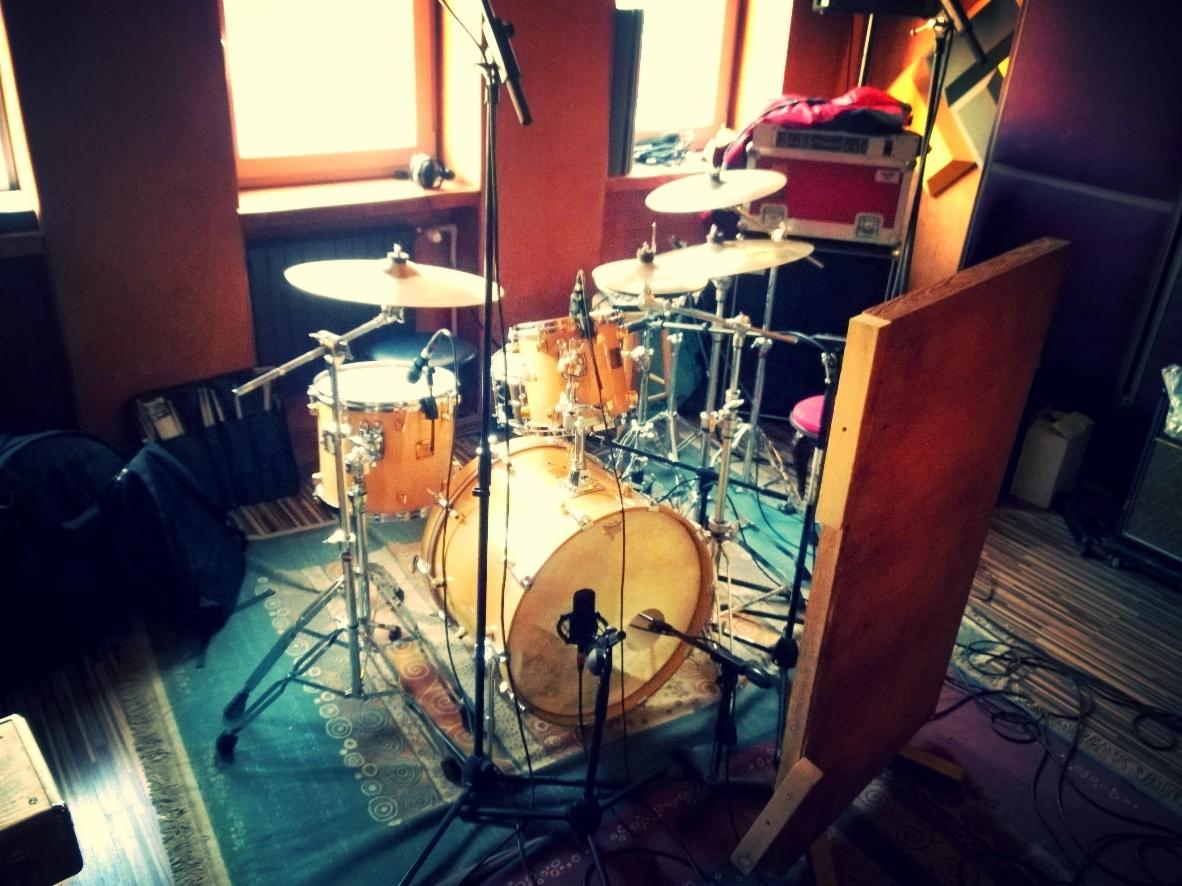 Zestaw perkusyjny w pomarańczowym pomieszczeniu, z mikrofonami i panelami akustycznymi. Widoczne okno z naturalnym światłem i dywan z wzorem.