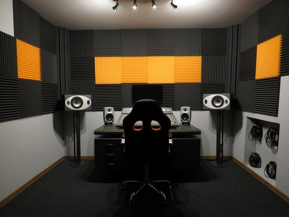 Profesjonalne studio nagrań z panelami akustycznymi w odcieniach szarości i pomarańczy. Widoczne monitory studyjne, mikser i ergonomiczne krzesło gamingowe.