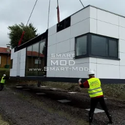 Montaż modułowego domu z białymi panelami elewacyjnymi za pomocą dźwigu. Widoczni pracownicy w odblaskowych kamizelkach i kaskach. Na budynku logo firmy.