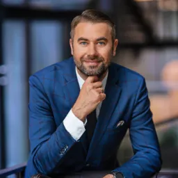 Portret uśmiechniętego doradcy w niebieskim garniturze w kratę, opierającego się o skórzane krzesło, Kalisz Pomorski. Widoczny zegarek na lewym nadgarstku.