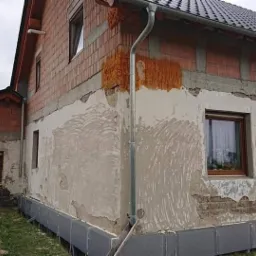 Dwukondygnacyjny dom w trakcie ocieplania elewacji: widoczne fragmenty ocieplenia na ścianie szczytowej, surowa cegła na g&oacute;rnej kondygnacji, częściowo otynkowana ściana z widocznymi śladami po...