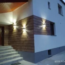 Dwukondygnacyjny dom z oświetloną elewacją imitującą drewno, widoczne schody wejściowe i fragment chodnika z kostki.