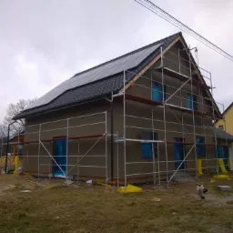Dom jednorodzinny w trakcie tynkowania elewacji, z zamontowanymi panelami słonecznymi na dachu i rusztowaniem wokół budynku, zabezpieczone okna folią. Widoczny pracownik na zewnątrz.