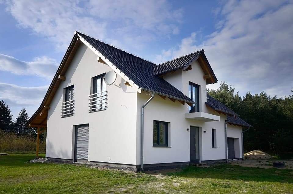 Nowoczesny dom jednorodzinny z białą elewacją, ciemną dachówką i grafitowymi oknami, widoczny z perspektywy ogrodu w słoneczny dzień.