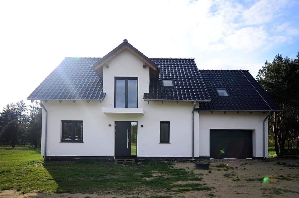 Nowoczesny dom jednorodzinny z białą elewacją, grafitową stolarką okienną i drzwiową, oraz ciemną dachówką, na tle zieleni i błękitnego nieba.