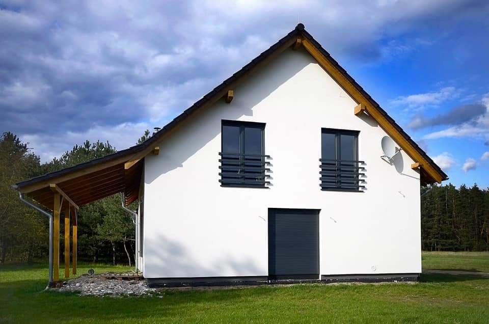 Nowoczesny dom jednorodzinny z białą elewacją, ciemnymi oknami z balkonami francuskimi i drewnianym zadaszeniem nad wejściem bocznym, antena satelitarna na ścianie szczytowej.