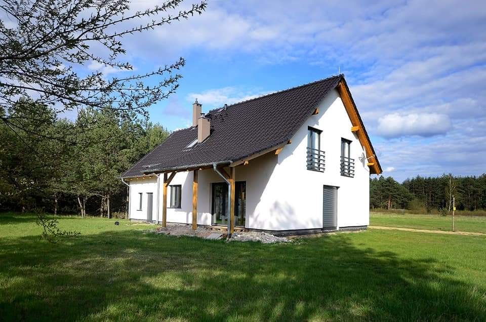 Nowoczesny dom jednorodzinny z białą elewacją, ciemną dachówką i drewnianymi elementami werandy, otoczony zielonym trawnikiem i lasem w tle, uchwycony w słoneczny dzień.