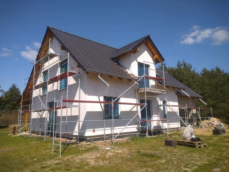 Dom jednorodzinny w trakcie prac elewacyjnych, z widocznymi rusztowaniami i fragmentem ocieplonej ściany w szarym kolorze, na tle błękitnego nieba i drzew.