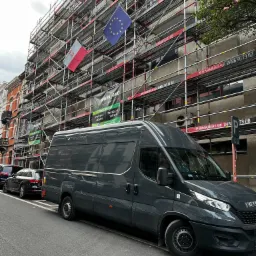 Szary bus firmy budowlanej zaparkowany przed budynkiem w rusztowaniach z flagami Polski i Unii Europejskiej, widoczne banery reklamowe na rusztowaniach.