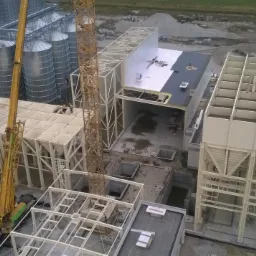 Budowa hali z użyciem dźwigu, widok z góry. Konstrukcja stalowa w trakcie montażu, widoczne elementy dachu i elewacji. Silosy w tle.